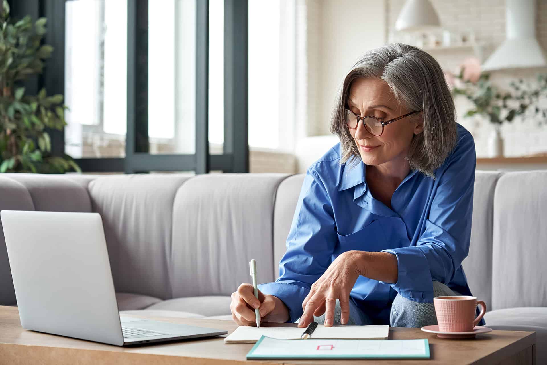 Eldre kvinne med briller sitter i en sofa ved en PC og et stuebord og noterer på papir